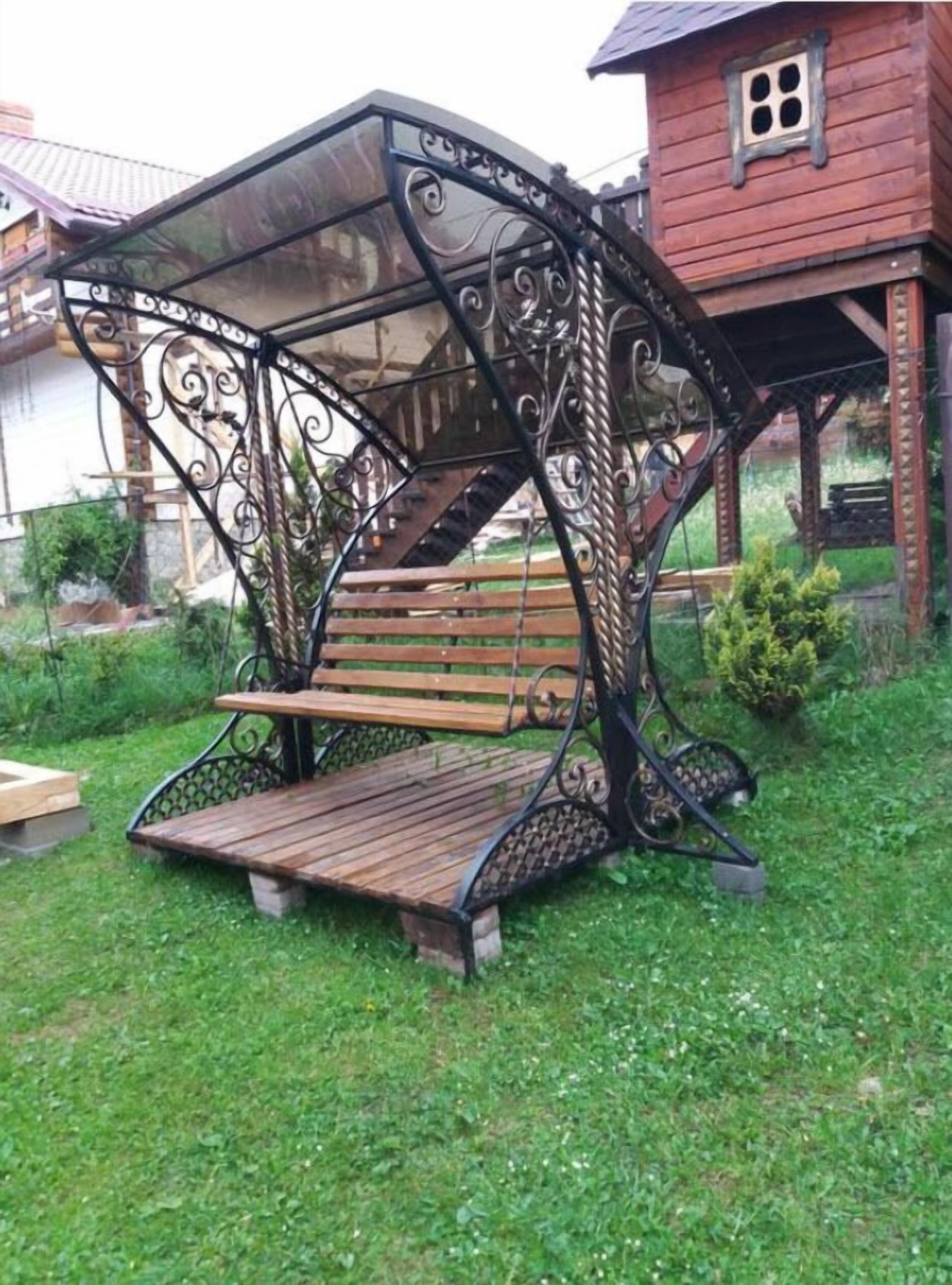 A wrought iron swing seat with a wooden bench, set on a grassy area with a house in the background.
