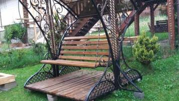 A wrought iron swing seat with a wooden bench, set on a grassy area with a house in the background.