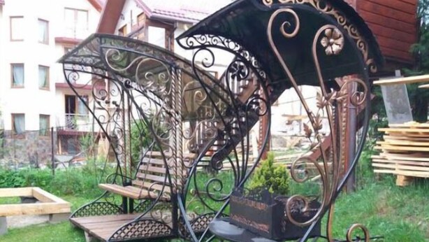A wrought iron bench with intricate designs, placed on a grassy area with a wooden bench and a planter nearby. - Saduba Perluna Bukovel (Bukovel)