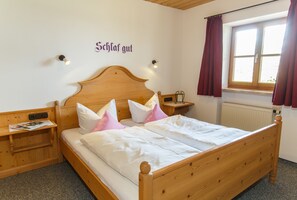 A wooden bed with white bedding, a nightstand with a lamp, and a window with curtains. - Ferienwohnungen Widdmoos (Ruhpolding)