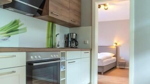 Une cuisine compacte avec des meubles blancs, un four encastré et un plan de travail en bois. On aperçoit un lit et une table de chevet dans la pièce adjacente.
