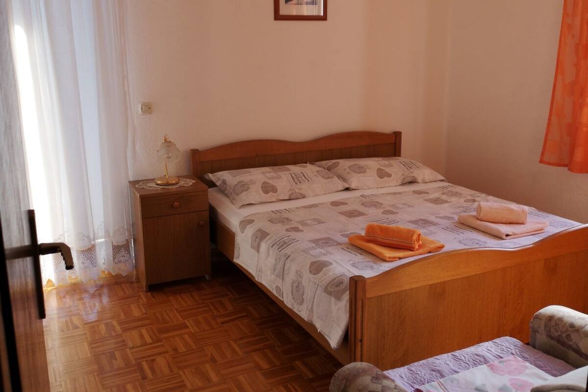 A bedroom with a wooden bed, a nightstand, a chair, and a window with sheer curtains.
