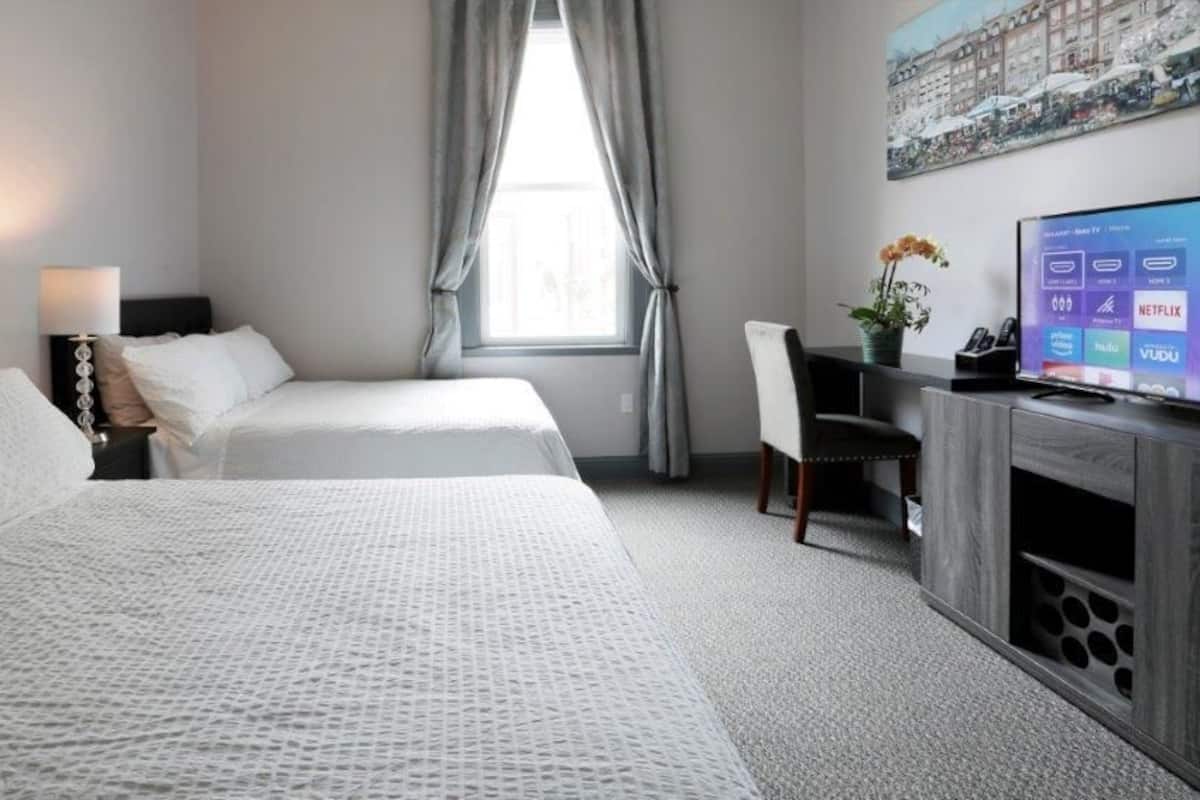 A hotel room with two beds, a desk with a computer, a TV displaying streaming services, and a window with curtains.