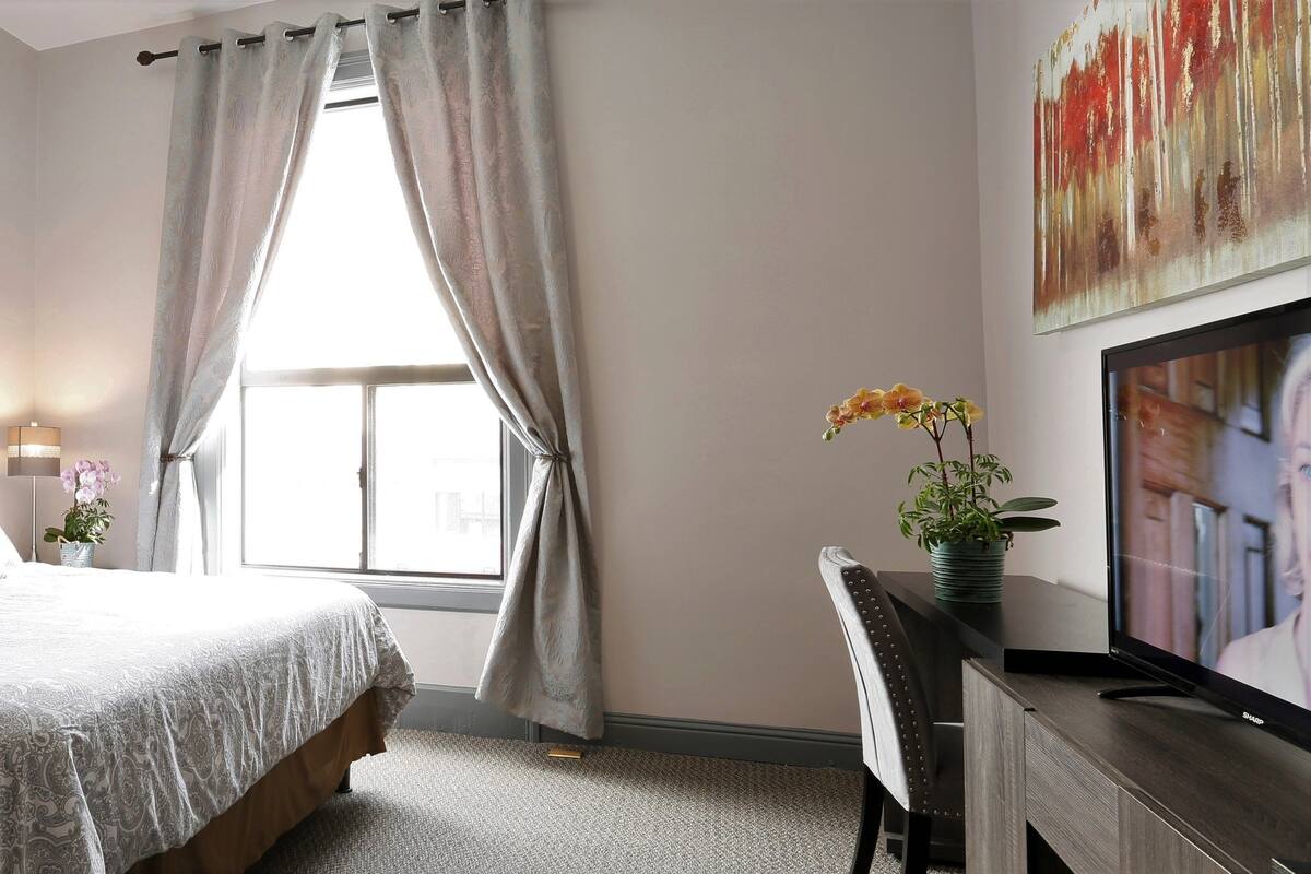 A bedroom with a tufted headboard, a large bed with patterned bedding, a desk with a computer, and a window with curtains.