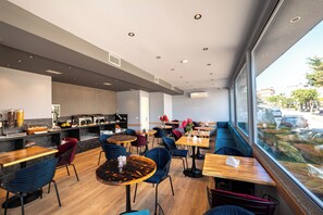 A modern cafe with wooden tables and chairs, a kitchen area, and large windows offering a view of the street. - Hotel Piramida (Ksamil)