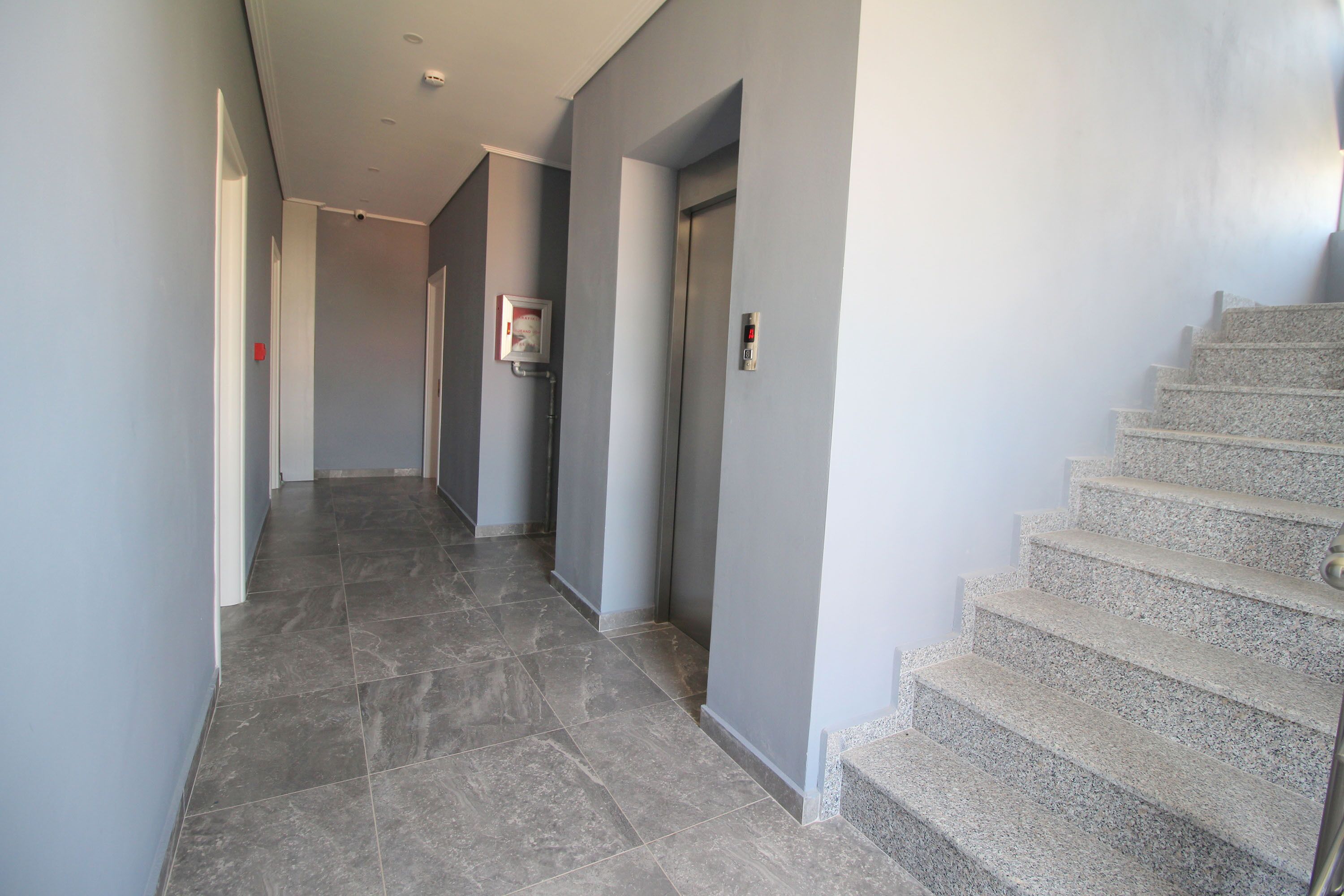 A modern hallway with a marble floor, a staircase on the right, and a door with a security camera on the left.