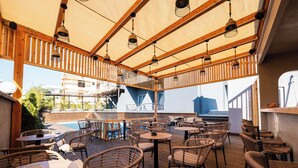 Una terraza al aire libre con mesas de madera y sillas de mimbre, con vista a una piscina y un edificio de fachada azul.