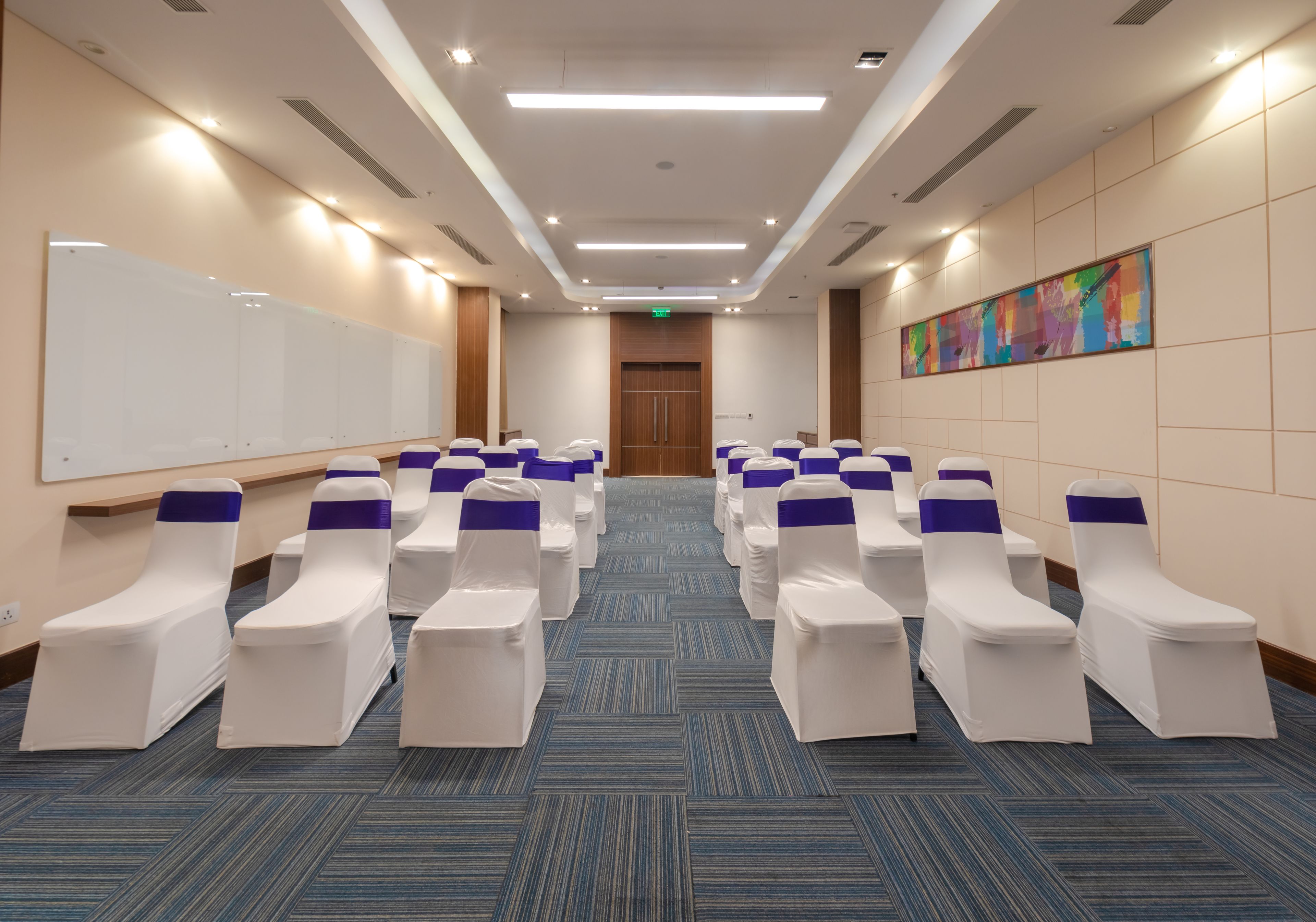 Une salle de conférence avec des rangées de chaises blanches ornées de rubans violets, un tableau blanc et une peinture abstraite aux couleurs vives accrochée au mur.