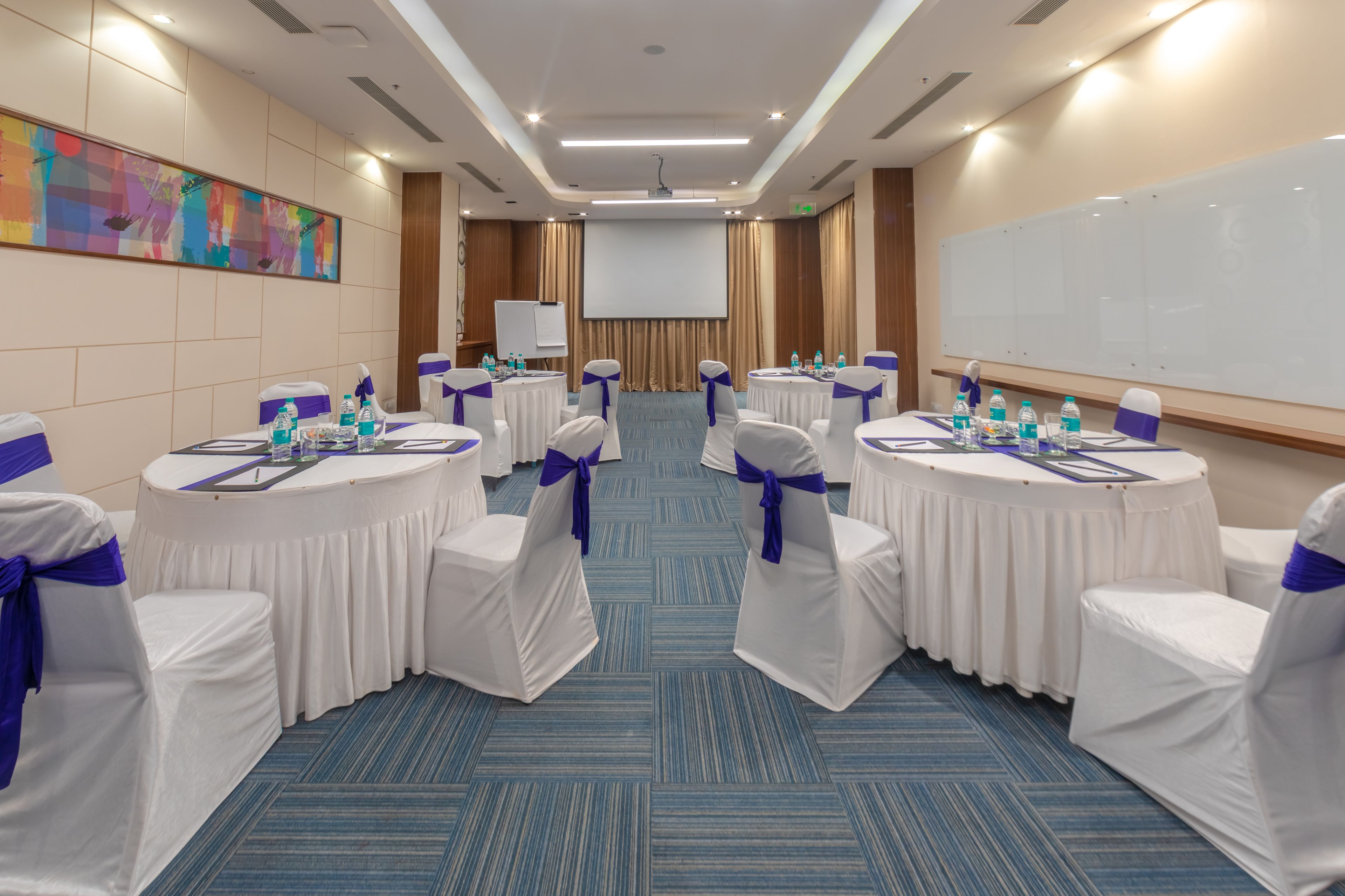 A conference room with round tables, white chairs with blue sashes, a projector screen, and a large whiteboard.