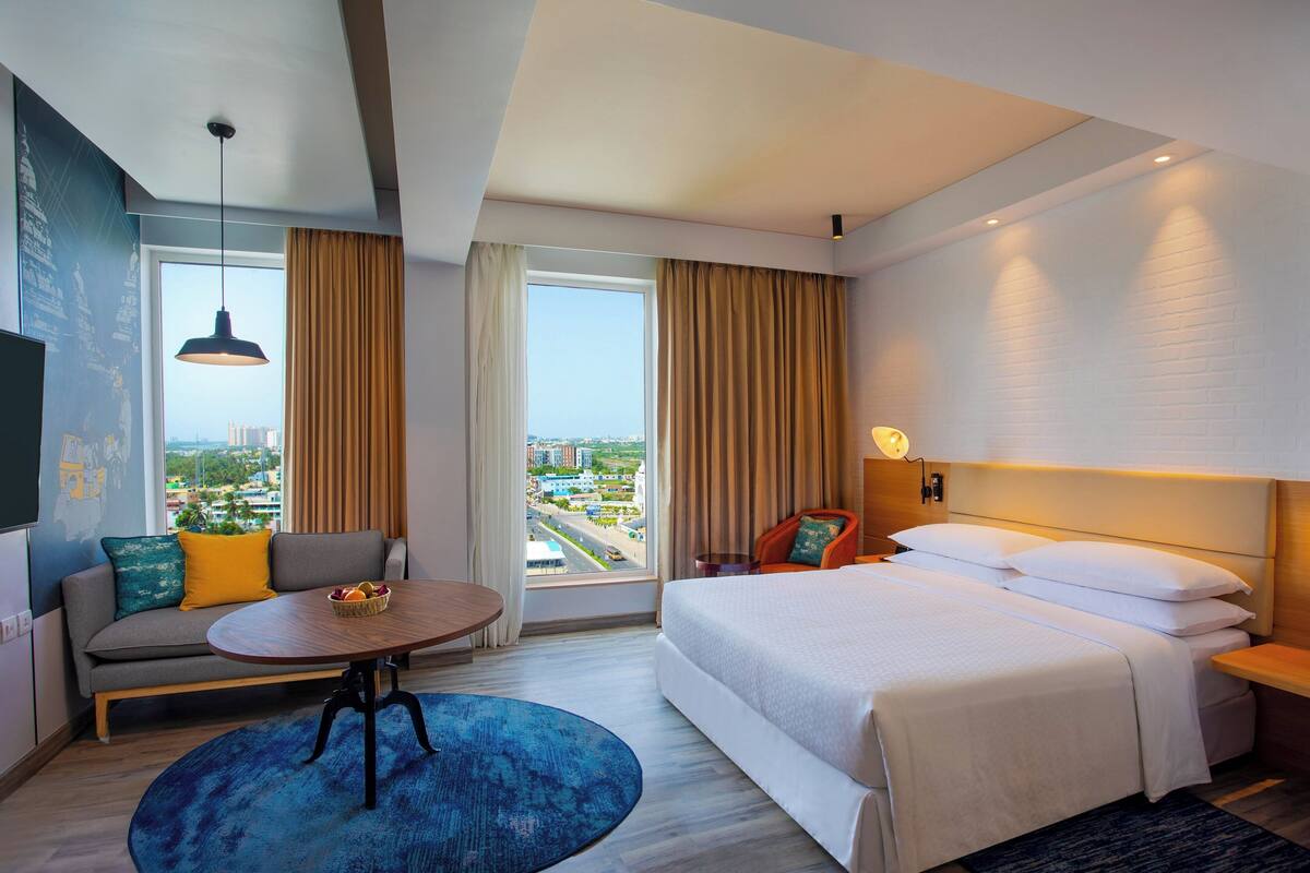 A hotel room with a bed, a sofa, a small table, a TV, and a view of the city.