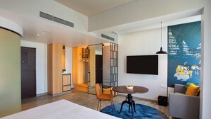 Una habitación de hotel moderna con una cama, una mesa de comedor redonda, un sofá, un televisor montado en la pared y un cartel grande.