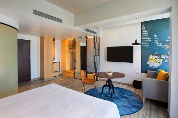 Una habitación de hotel moderna con una cama, una mesa de comedor redonda, un sofá, un televisor montado en la pared y un cartel grande.