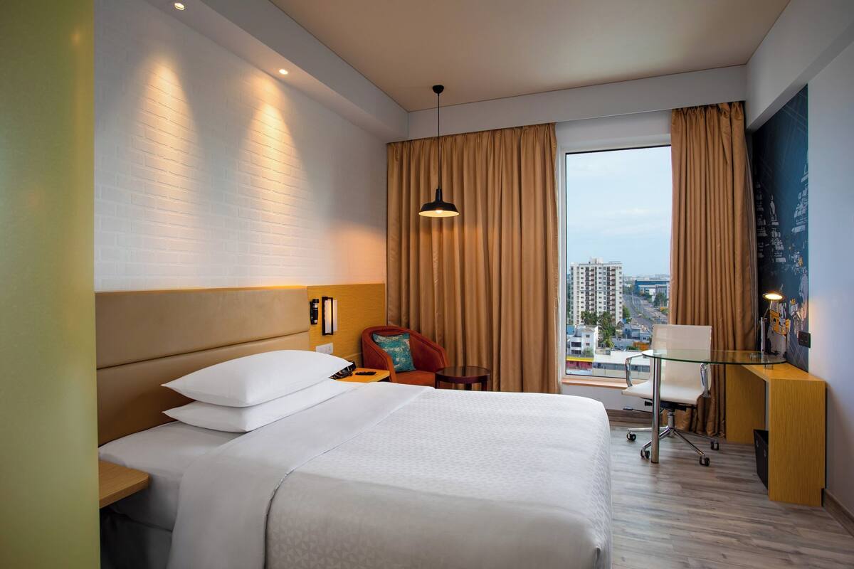 A hotel room with a large bed, a desk with a chair, a small table, and a view of the cityscape through the window.