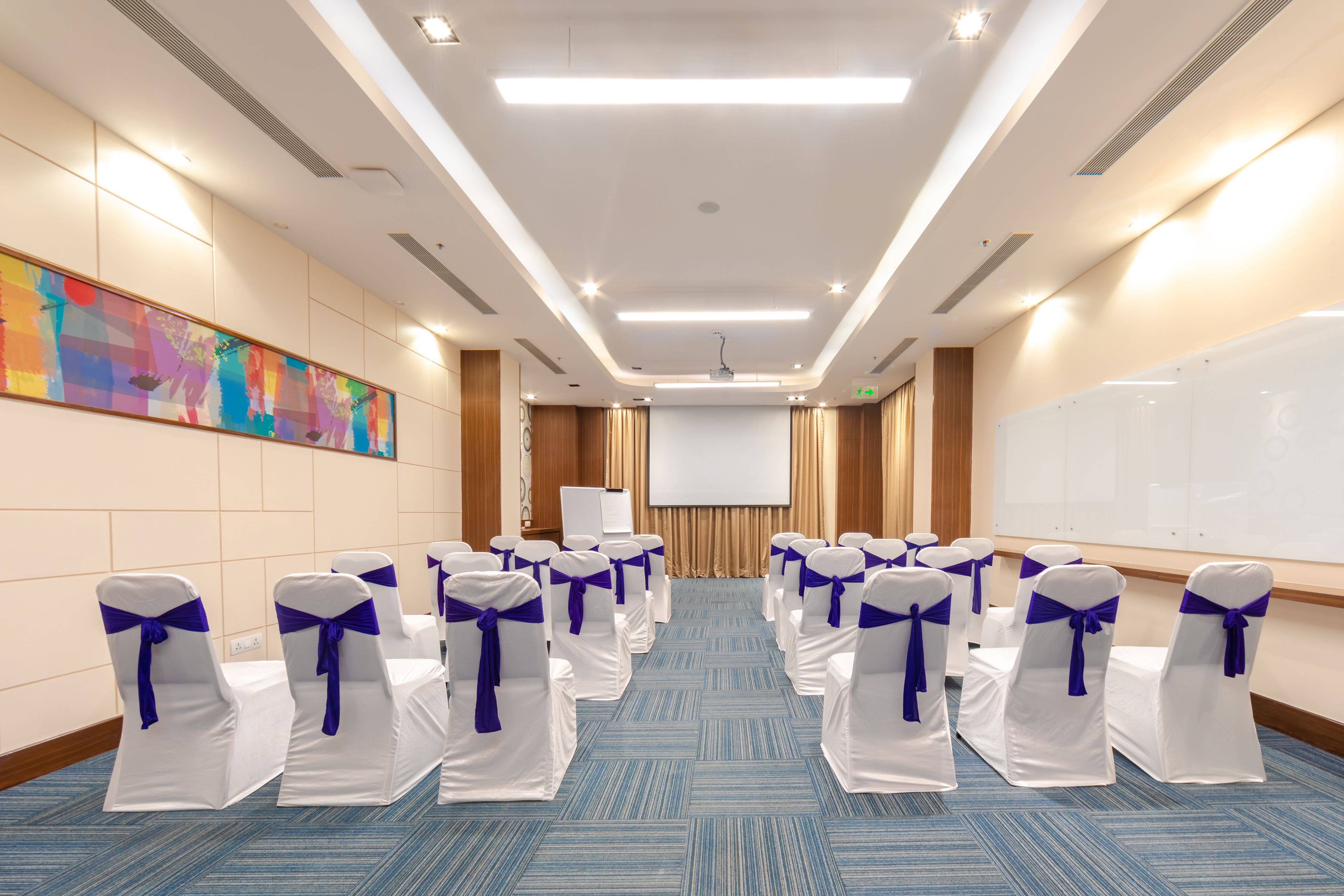 A conference room with rows of white chairs with blue sashes, a whiteboard, and a projector screen.