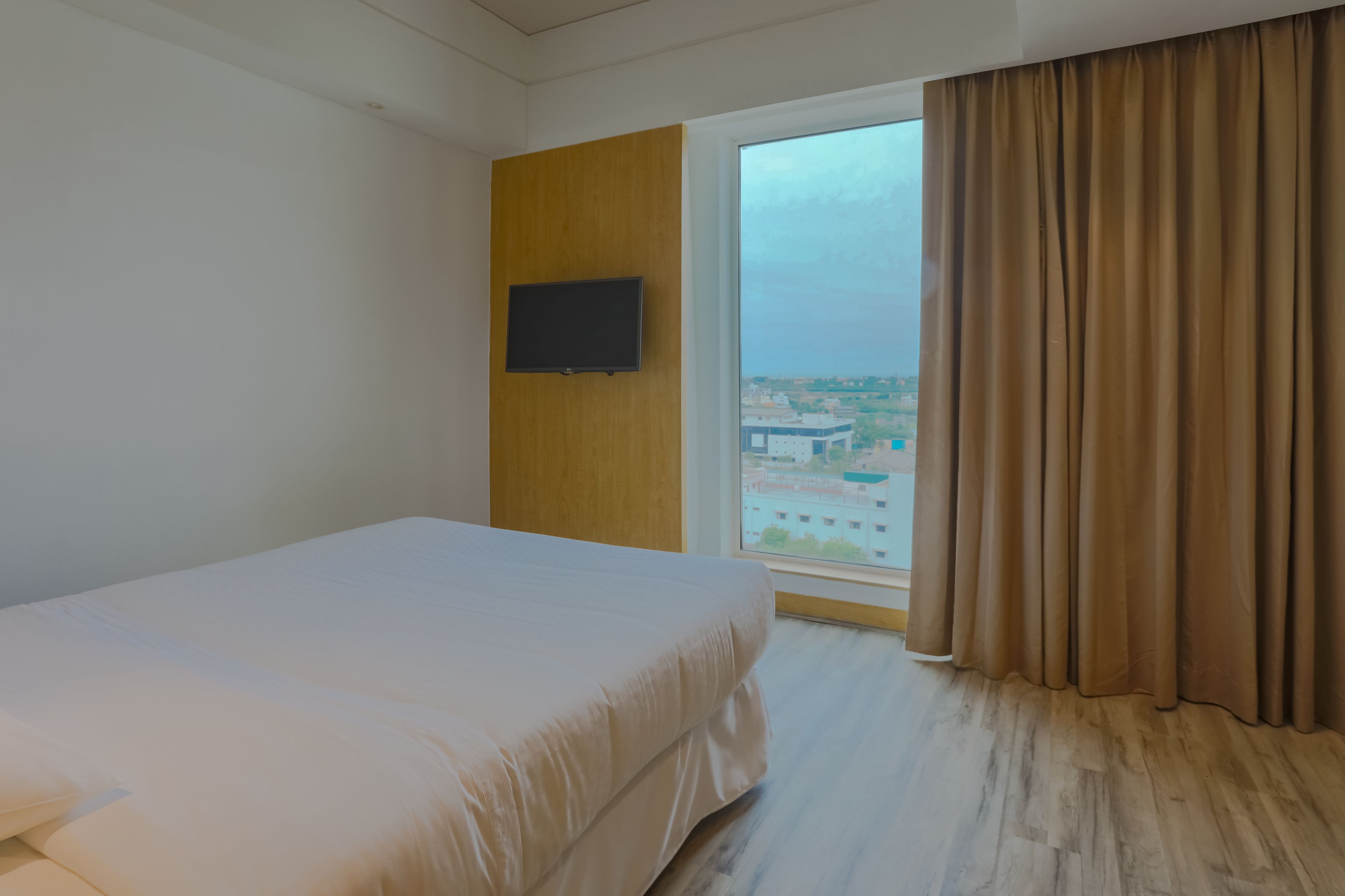 A hotel room with a bed, a TV mounted on a wooden panel, and a large window with curtains.