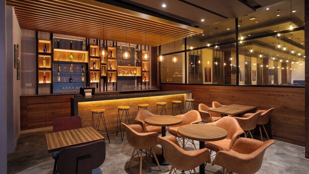 Un área de bar moderna con estantes de madera que exhiben botellas y vasos, y varios conjuntos de asientos con mesas y sillas redondas.