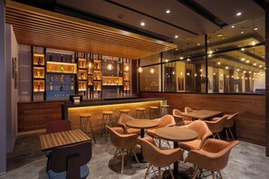 Un área de bar moderna con estantes de madera que exhiben botellas y vasos, y varios conjuntos de asientos con mesas y sillas redondas.