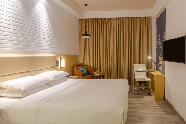 Habitación de hotel con una cama grande, un escritorio con una silla, una lámpara y un televisor en la pared.