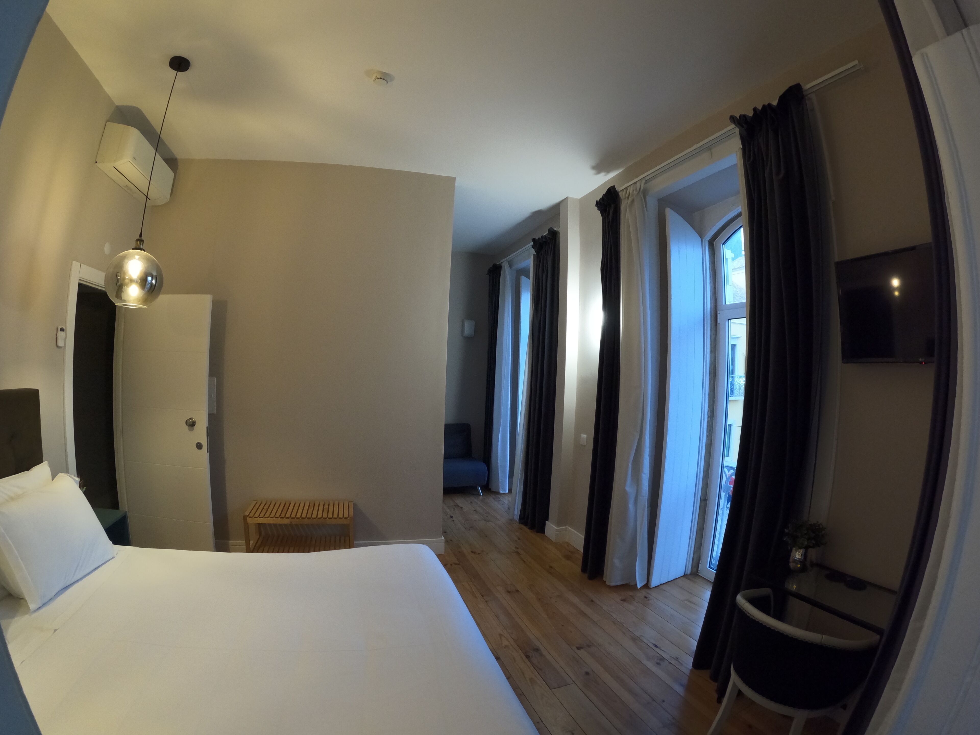 A hotel room with a large bed, a wooden bench, a small table, a TV, and a view of the outside through the curtains.