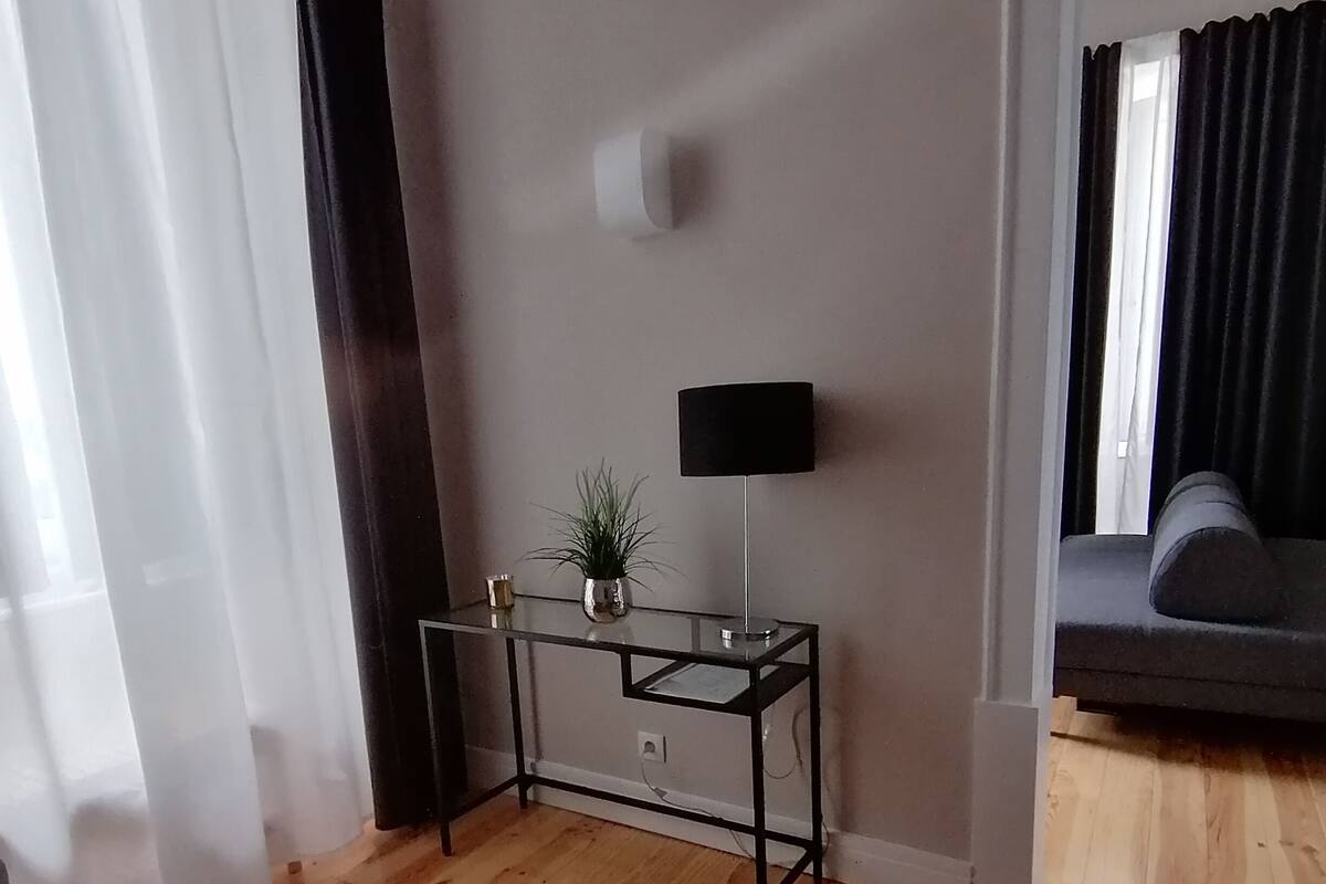 A room with a wooden floor, a glass table with a plant, a black lamp, and a grey sofa.