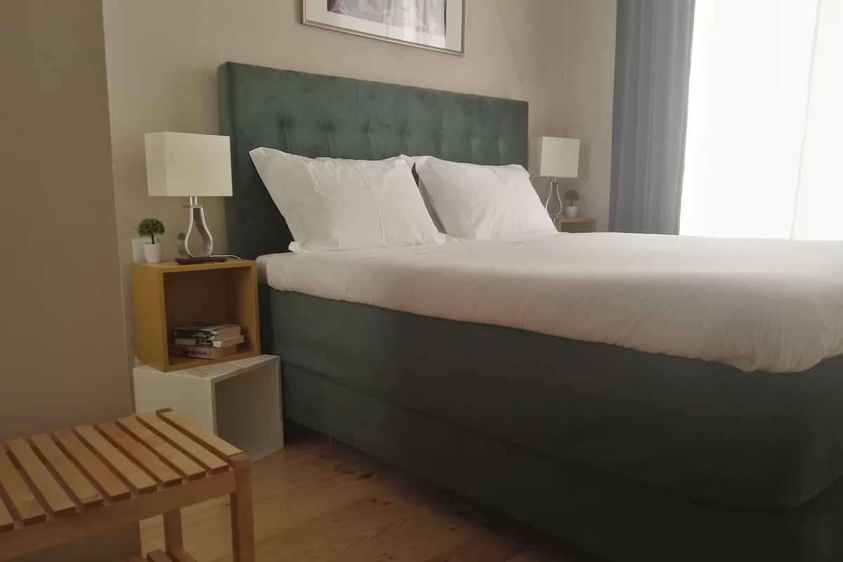 A neatly arranged hotel room with a large bed, two bedside tables with lamps, a small shelf with books, and a wooden bench.