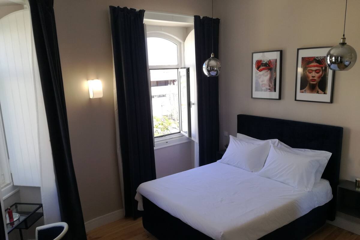 A hotel room with a bed, a chair, a desk, a window with curtains, and two wall-mounted pictures.