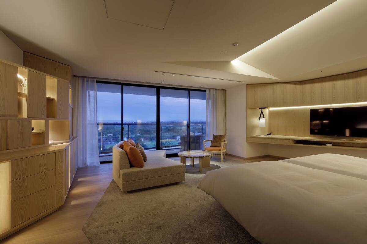 A modern hotel room with a large bed, a flat-screen TV mounted on the wall, a seating area with a sofa and armchair, and a balcony with a city view.