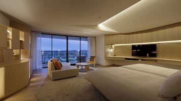 Une chambre d’hôtel moderne dotée d’un grand lit, d’un téléviseur à écran plat fixé au mur, d’un coin salon avec un canapé et un fauteuil, et d’un balcon offrant une vue sur la ville.