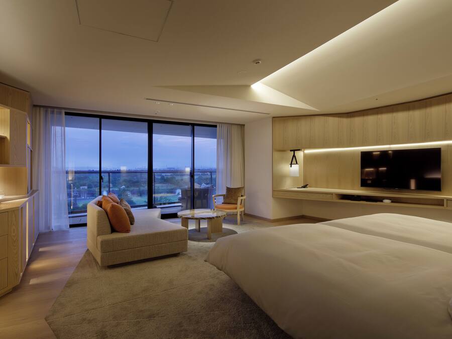 Une chambre d’hôtel moderne dotée d’un grand lit, d’un téléviseur à écran plat fixé au mur, d’un coin salon avec un canapé et un fauteuil, et d’un balcon offrant une vue sur la ville.