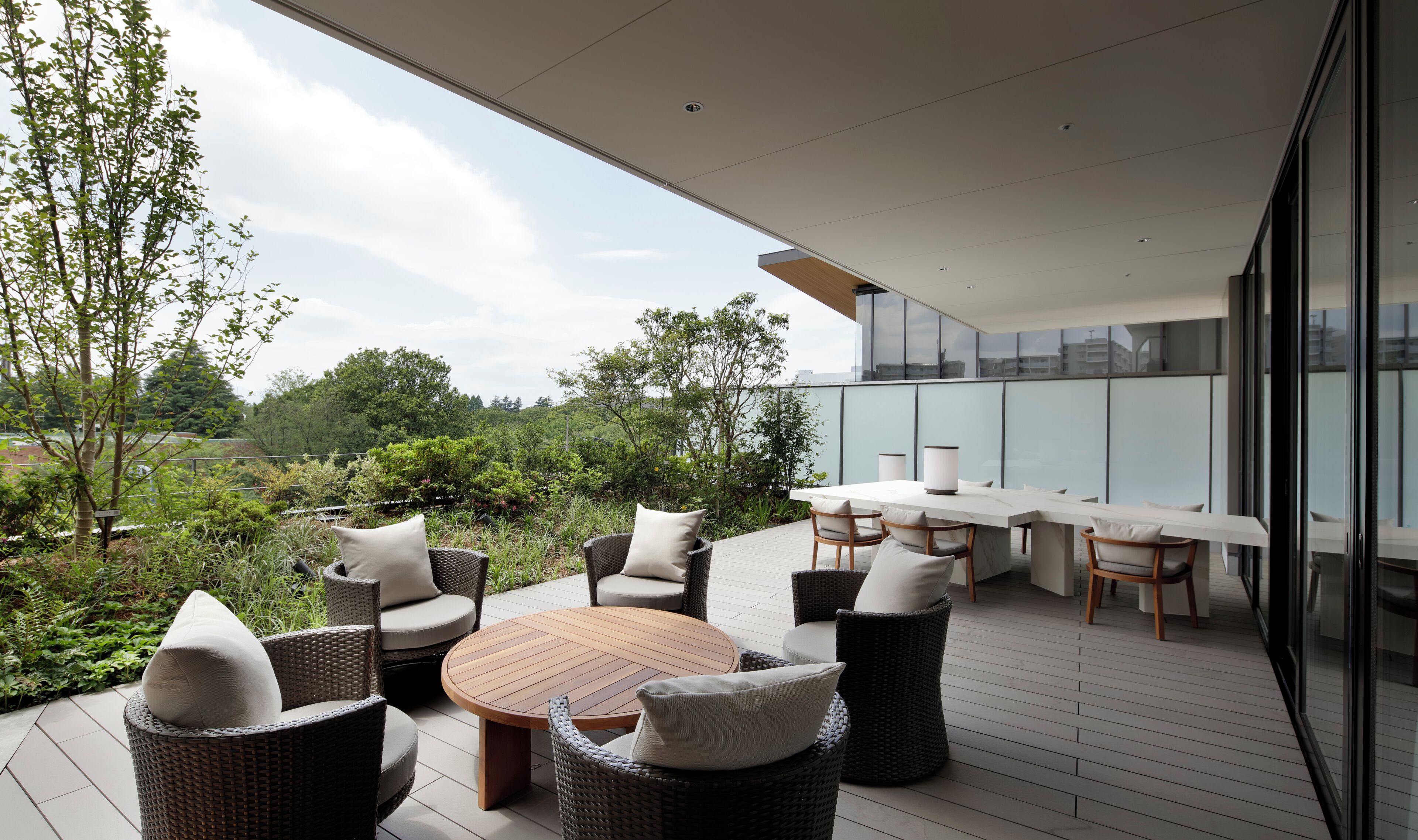 A modern outdoor patio with wicker furniture, a round wooden table, and a glass balustrade overlooking a garden.
