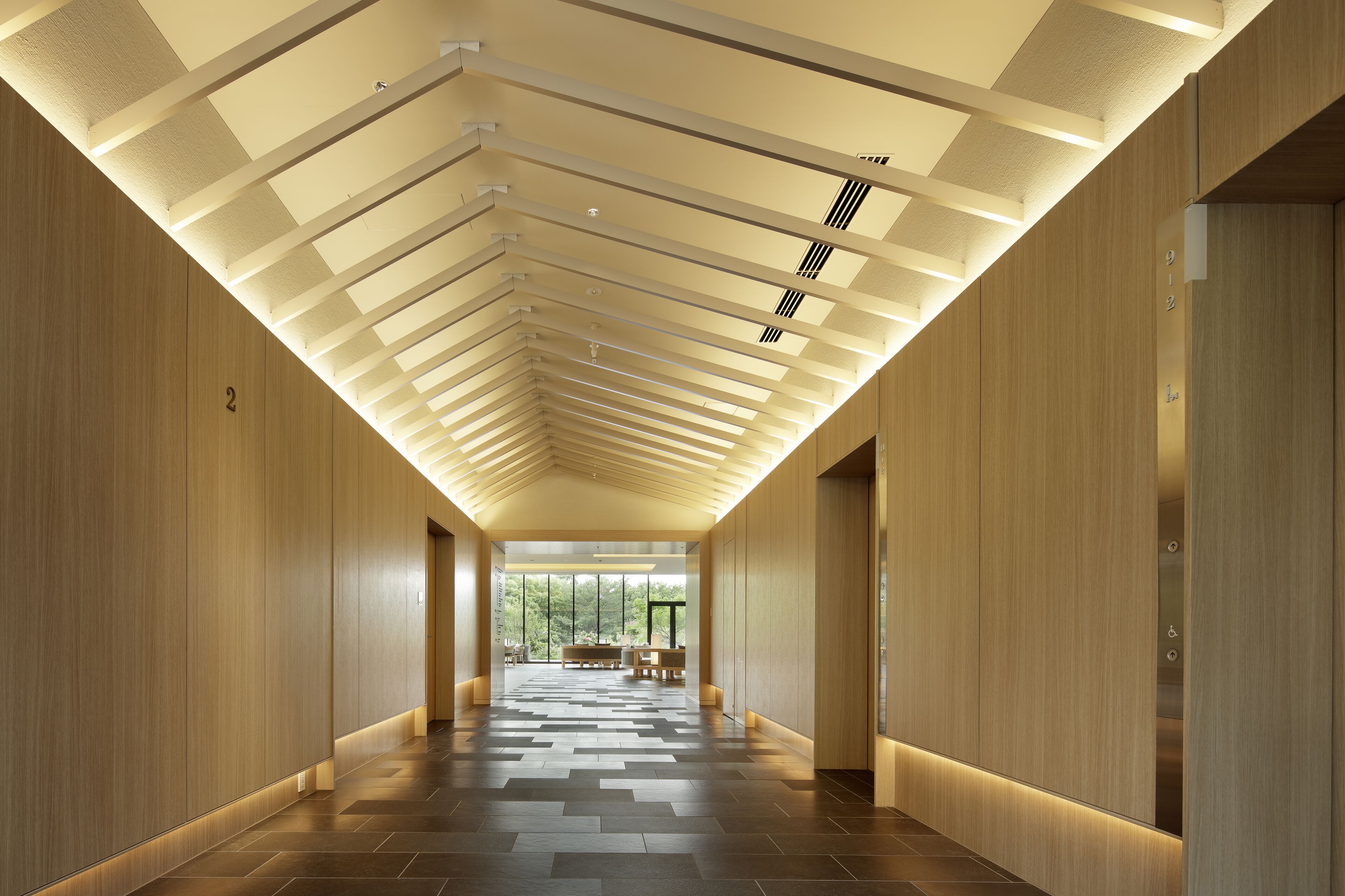 A modern hallway with a wooden floor and ceiling, featuring a bench and a view of greenery outside.