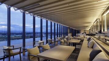 Un restaurant moderne doté de grandes fenêtres offrant une vue sur la ville, de hauts plafonds avec un éclairage linéaire et proposant différents types de sièges, notamment des tables et des canapés.