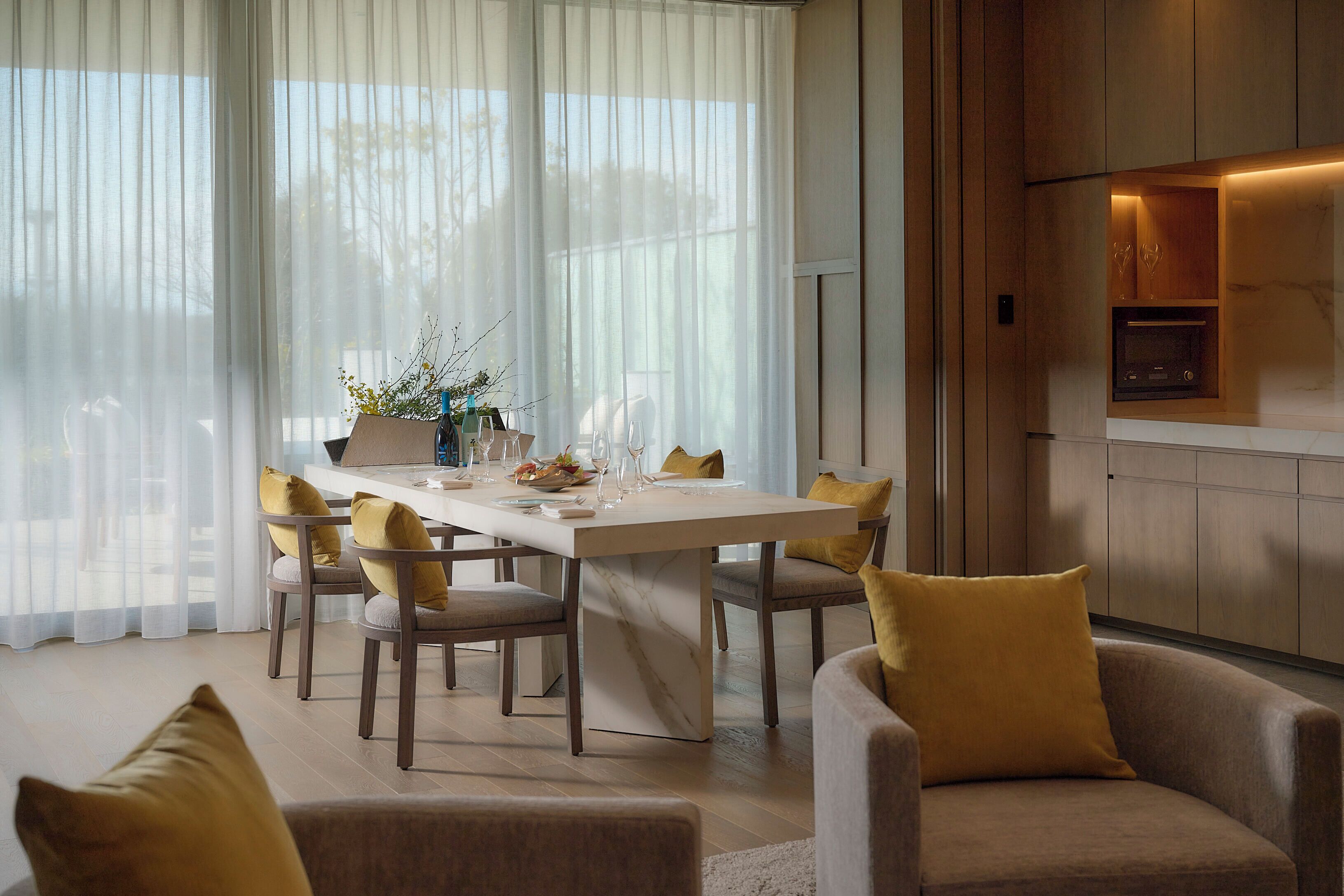 A dining area with a table set for four, chairs with cushions, a kitchen area with a microwave, and a large window with sheer curtains.