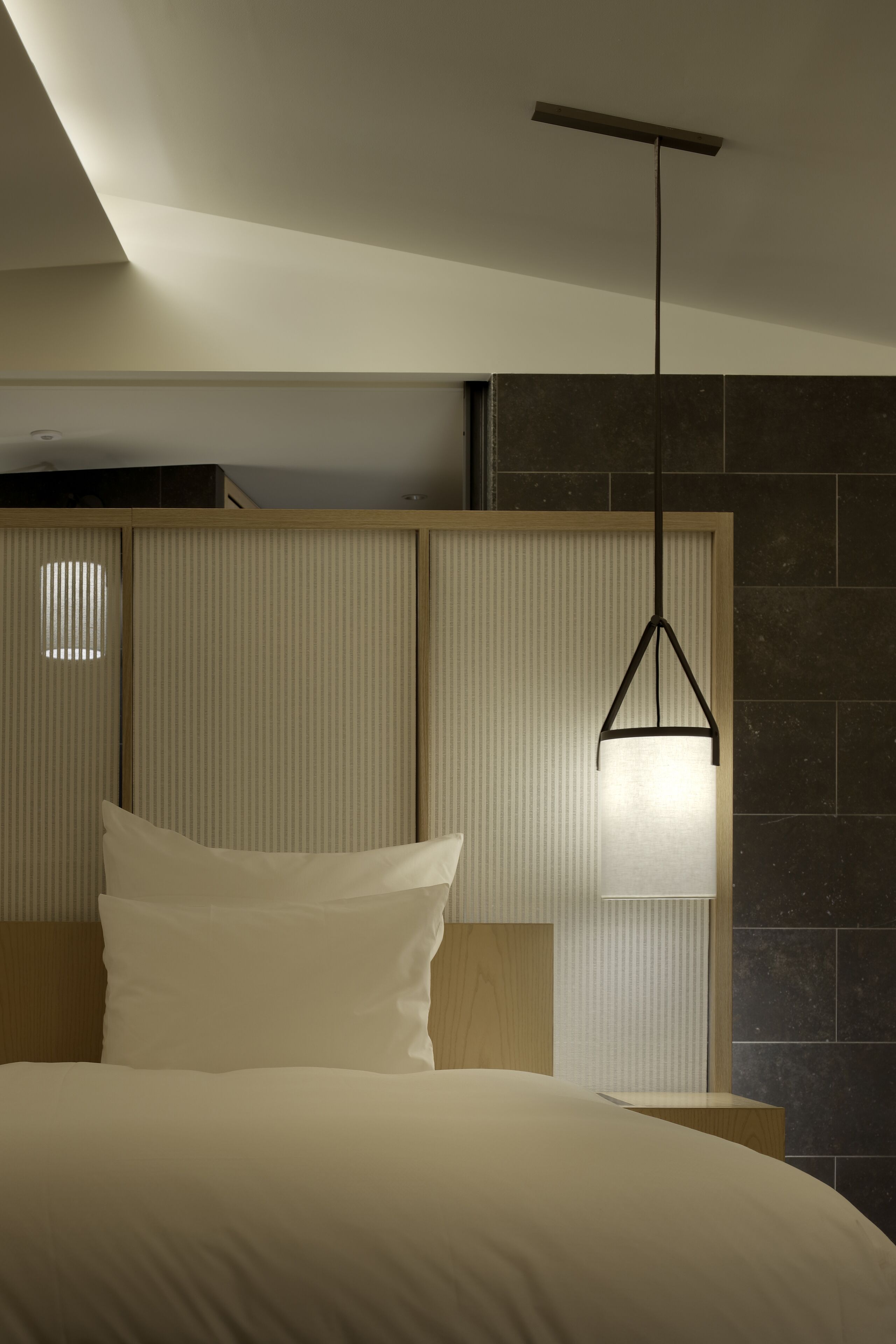 A modern hotel room with a bed, a bedside table, a hanging light fixture, and a textured wall panel.