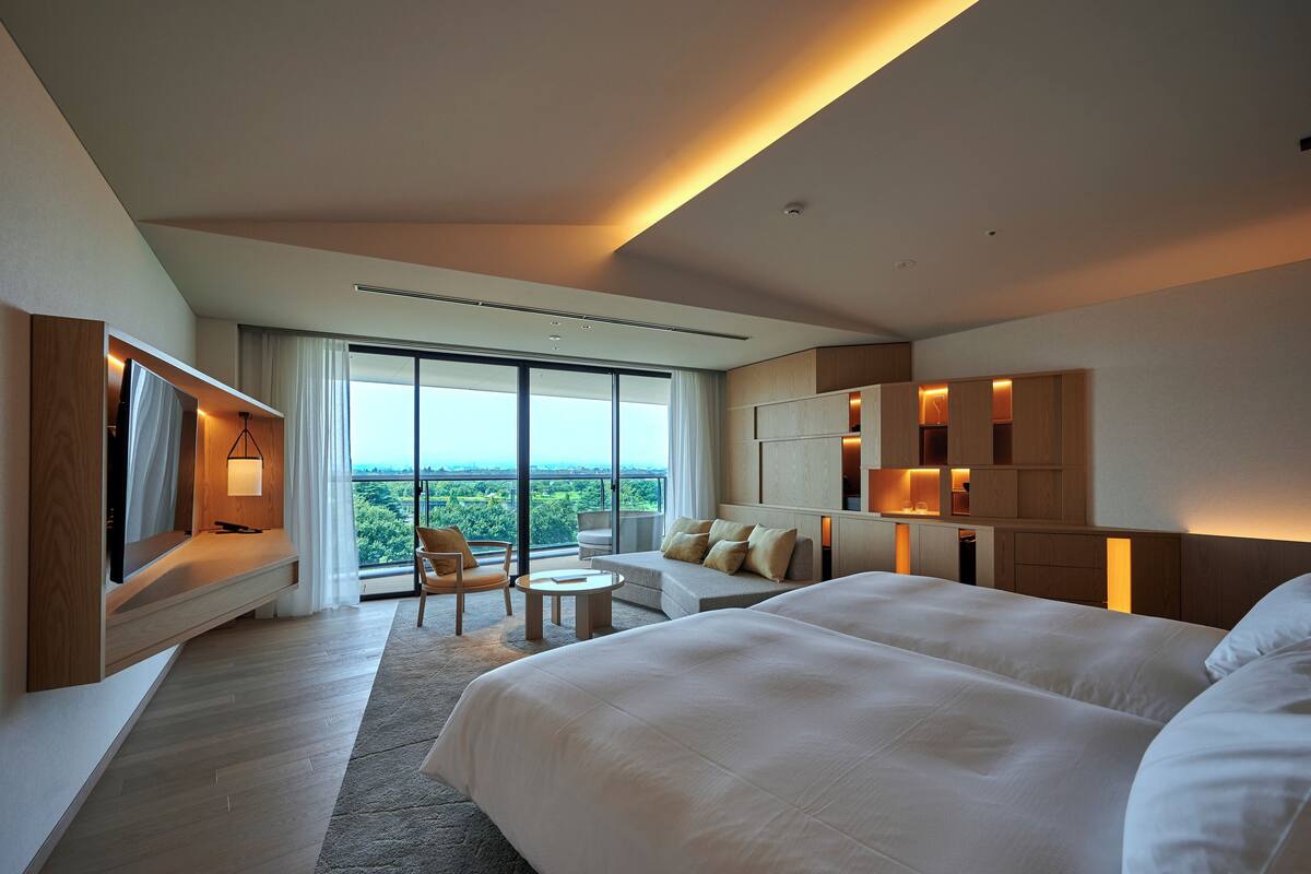 A modern hotel room with a large bed, a seating area with a table, a TV mounted on the wall, and a large window offering a view of the outdoors.