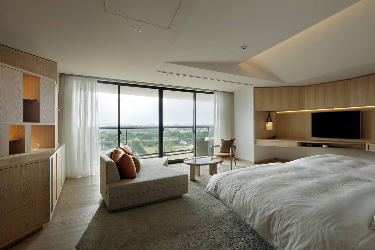 A modern hotel room with a large bed, a sofa, a TV, and a balcony with a city view.