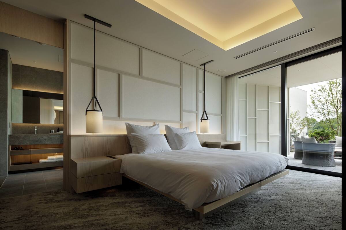 A modern bedroom with a large bed, bedside tables, a built-in bench, and a view through a large window.