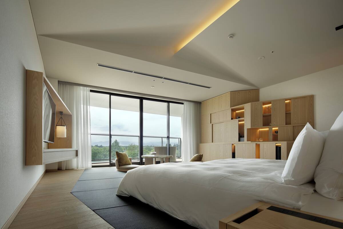 A modern hotel room with a large bed, built-in wooden shelving, a seating area, and a view of the outdoors.