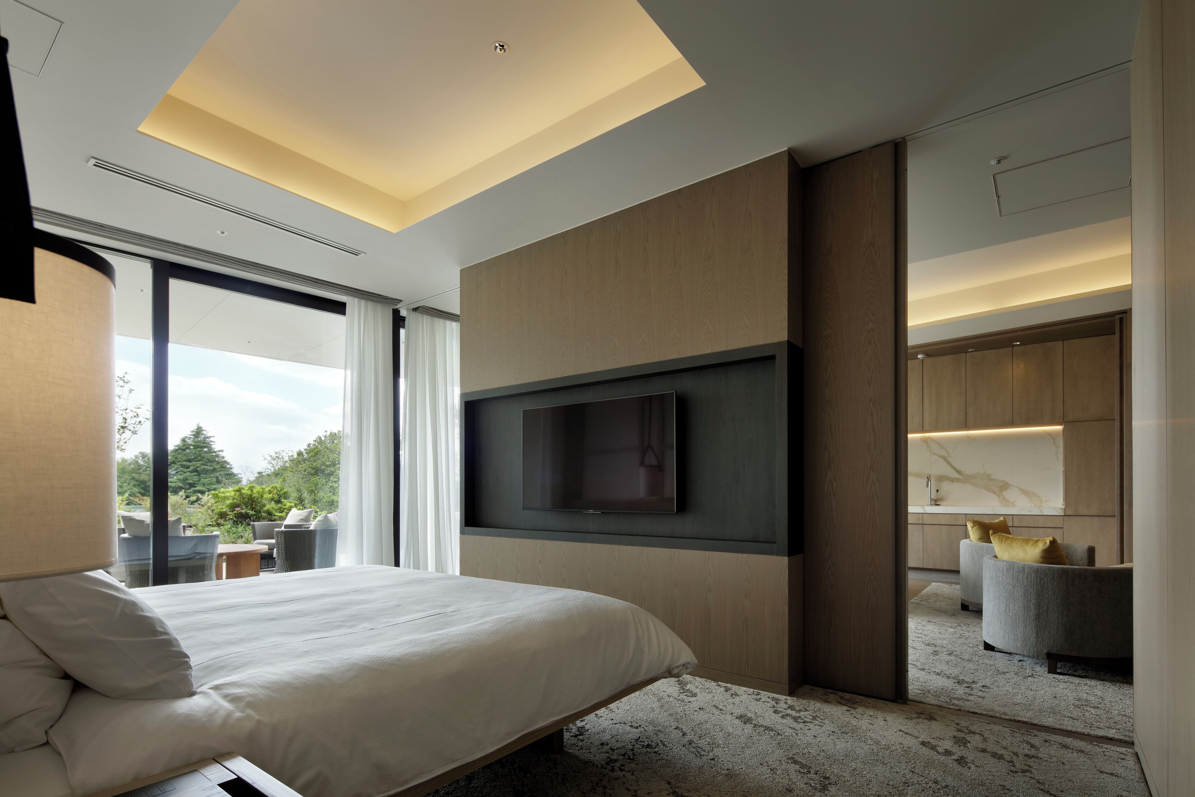 A modern hotel room with a large bed, a flat-screen TV mounted on the wall, a seating area with chairs, and a view of the outdoors.