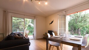 A modern living room with a sofa, dining table, and chairs. There is a TV on a stand and a balcony with a view of trees. - Soothing 4-person apartment in the heart of Erbalunga (Erbalunga)