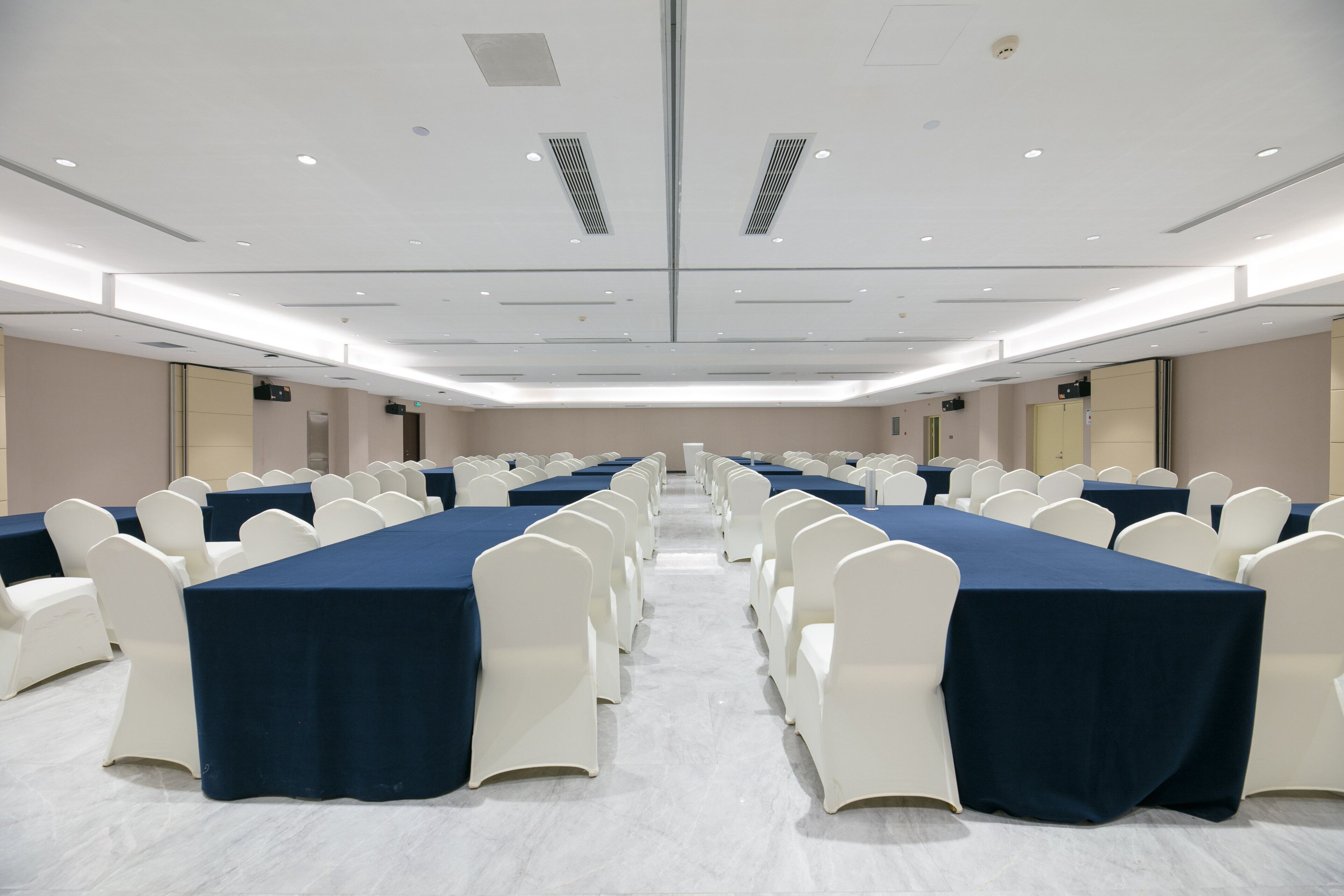 A conference room with rows of tables and chairs, equipped with ceiling lights and ventilation.