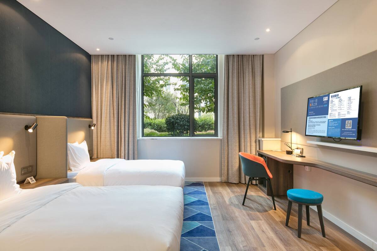 Habitación de hotel con dos camas, un escritorio, una silla, un televisor y una ventana con vistas a zonas verdes.