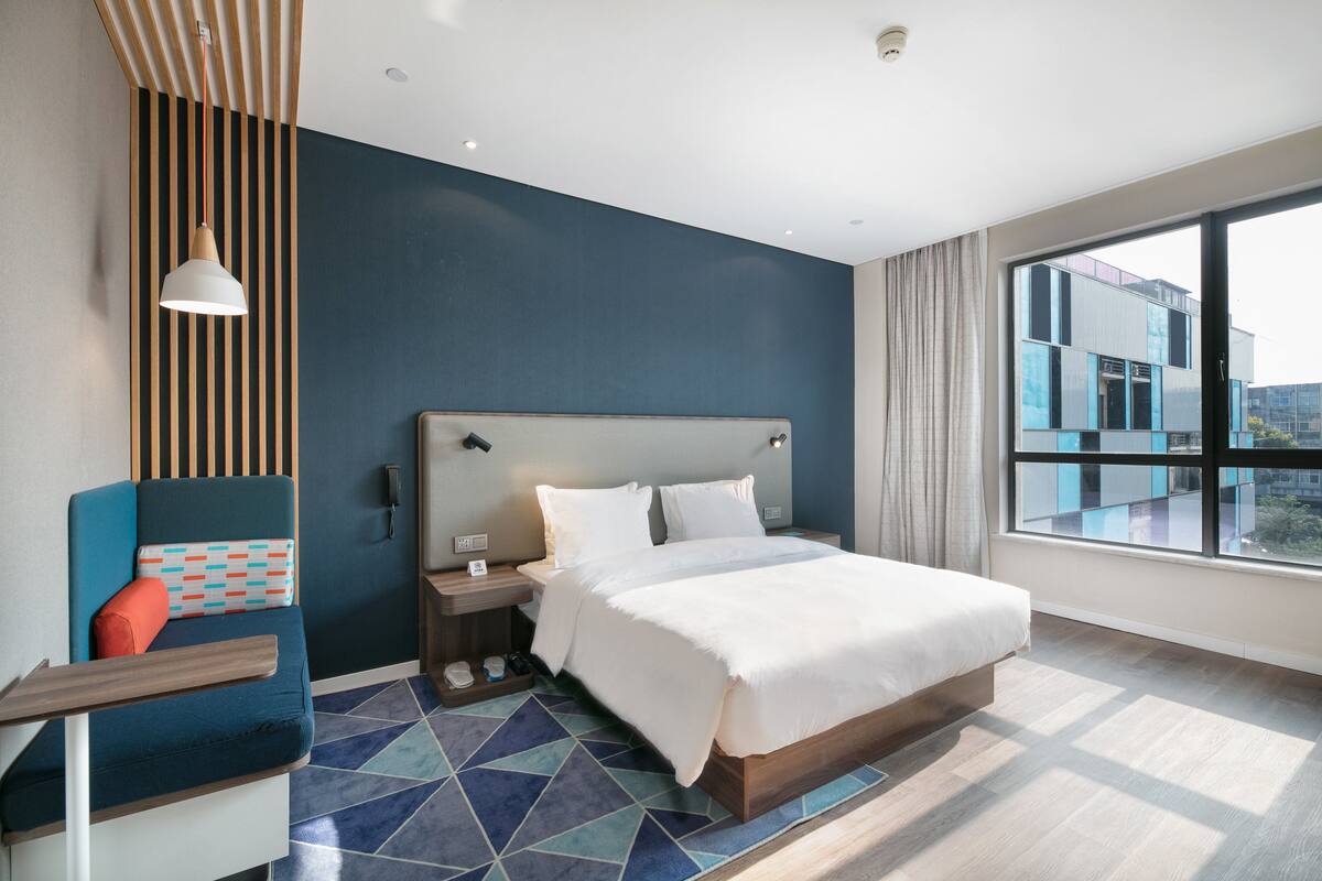 Una habitación de hotel moderna con una cama grande, un área de descanso con un cojín estampado, una mesita de noche y vistas a la ciudad a través de amplias ventanas.