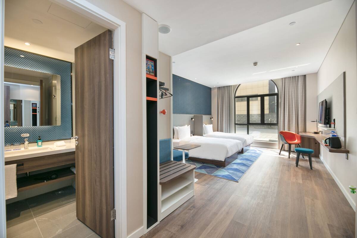 Una habitación de hotel moderna con una cama grande, un escritorio, una silla, un televisor y un baño con espejo y lavamanos.