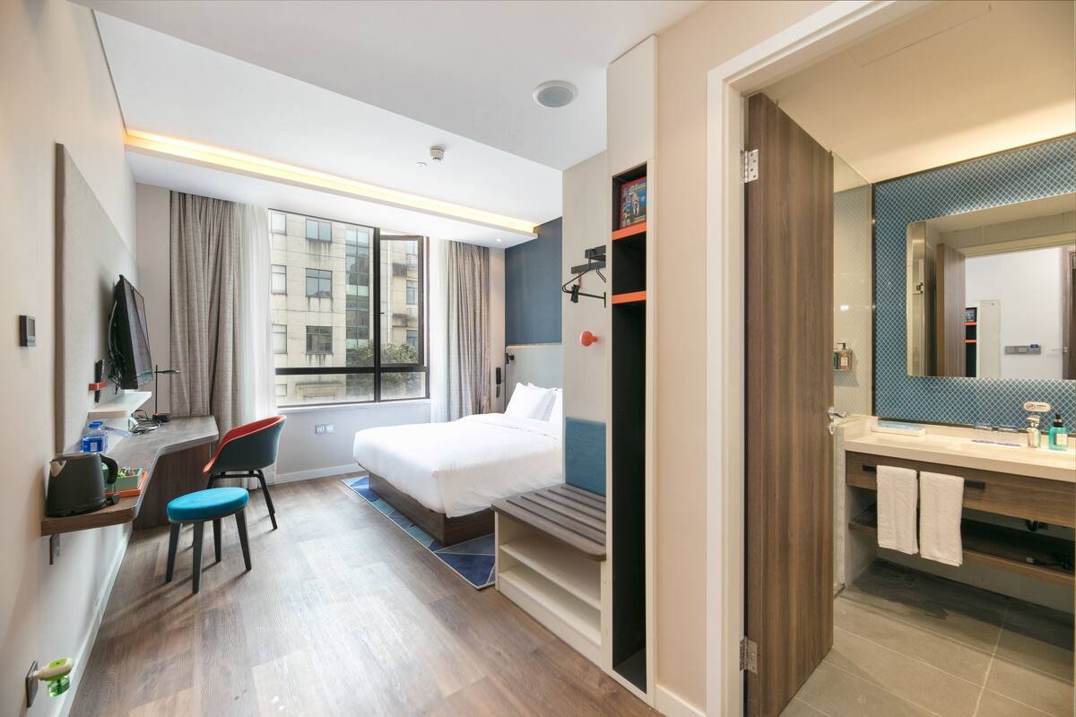 Habitación de hotel con cama, escritorio con silla, baño con espejo y ventana con cortinas.