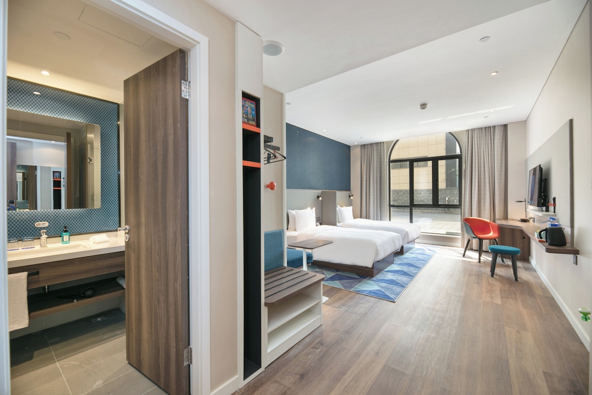 A hotel room with a large bed, a desk, a chair, a TV, and a bathroom with a mirror and sink.