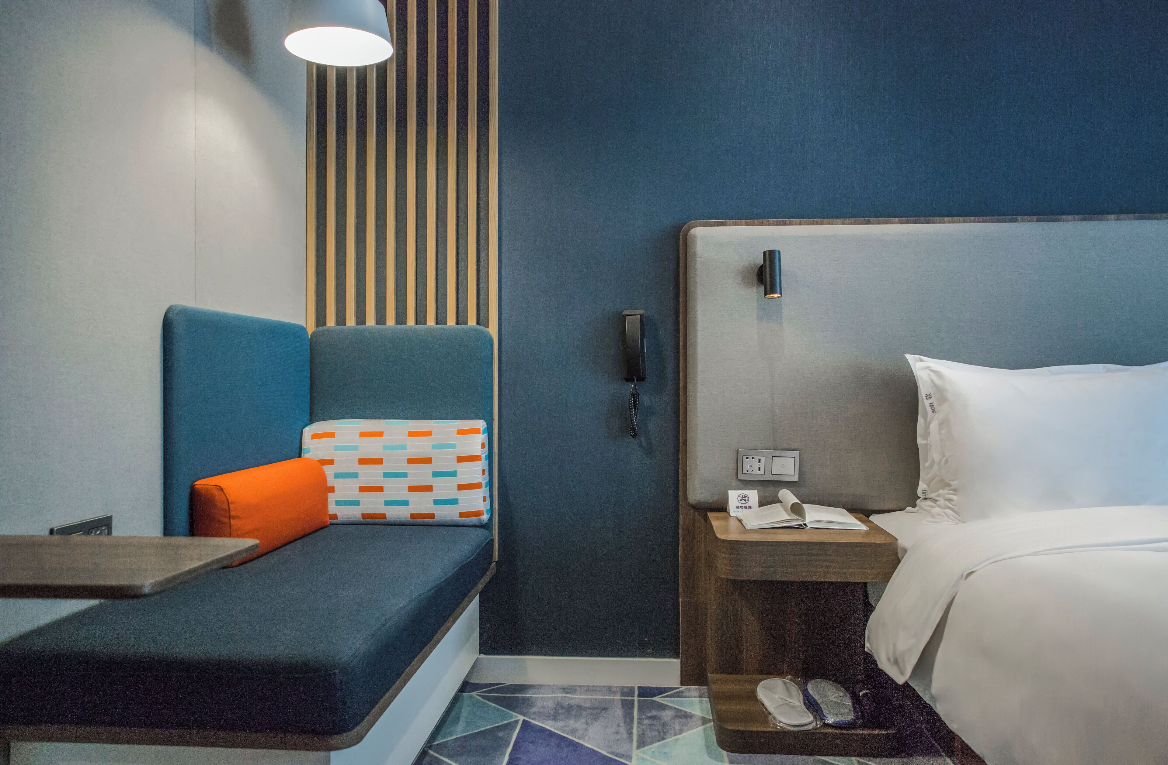 Ett modernt hotellrum med en blå vägg, en säng med vita sängkläder, en blå soffa med en orange kudde, en randig väggpanel och ett litet bord med en lampa.