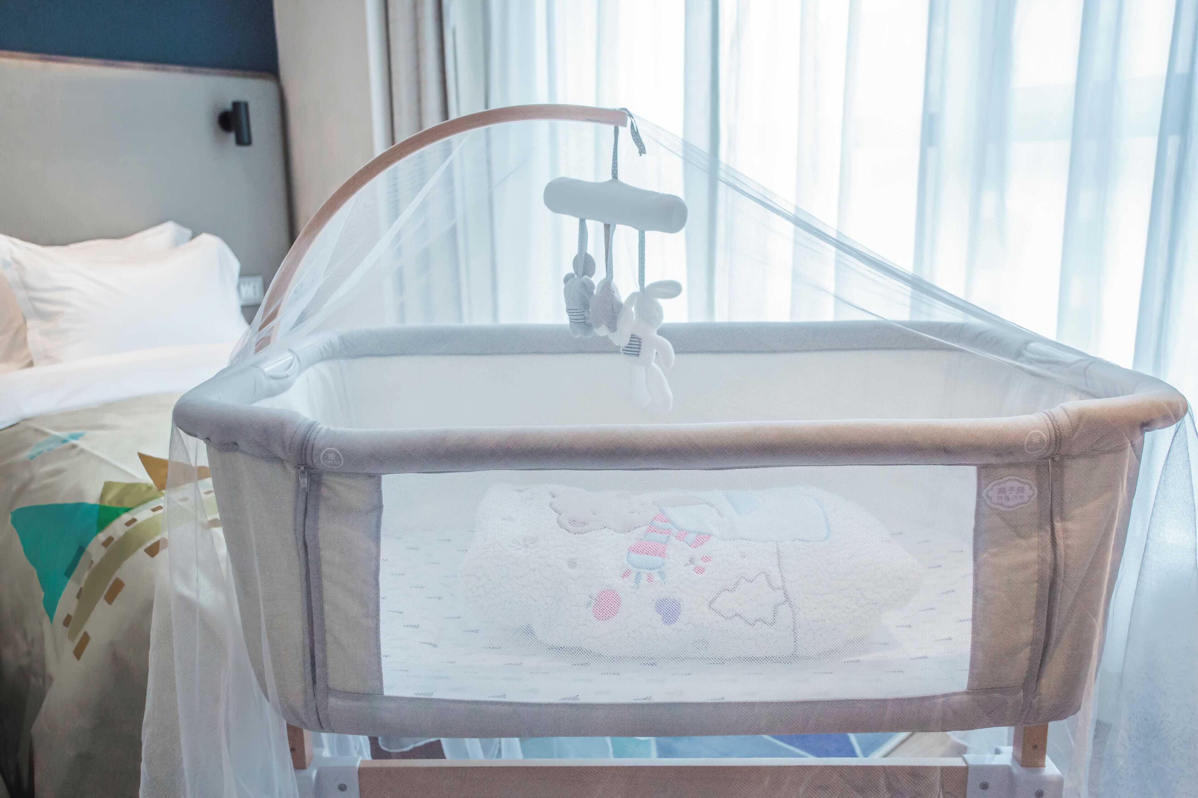 Una cuna para bebé con un móvil colgante, una ventana con cortinas translúcidas y una cama con ropa de cama blanca al fondo.