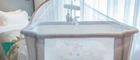 Una cuna para bebé con un móvil colgante, una ventana con cortinas translúcidas y una cama con ropa de cama blanca al fondo.