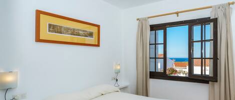 A hotel room with a bed, a framed picture on the wall, a window with curtains, and a view of the sea.